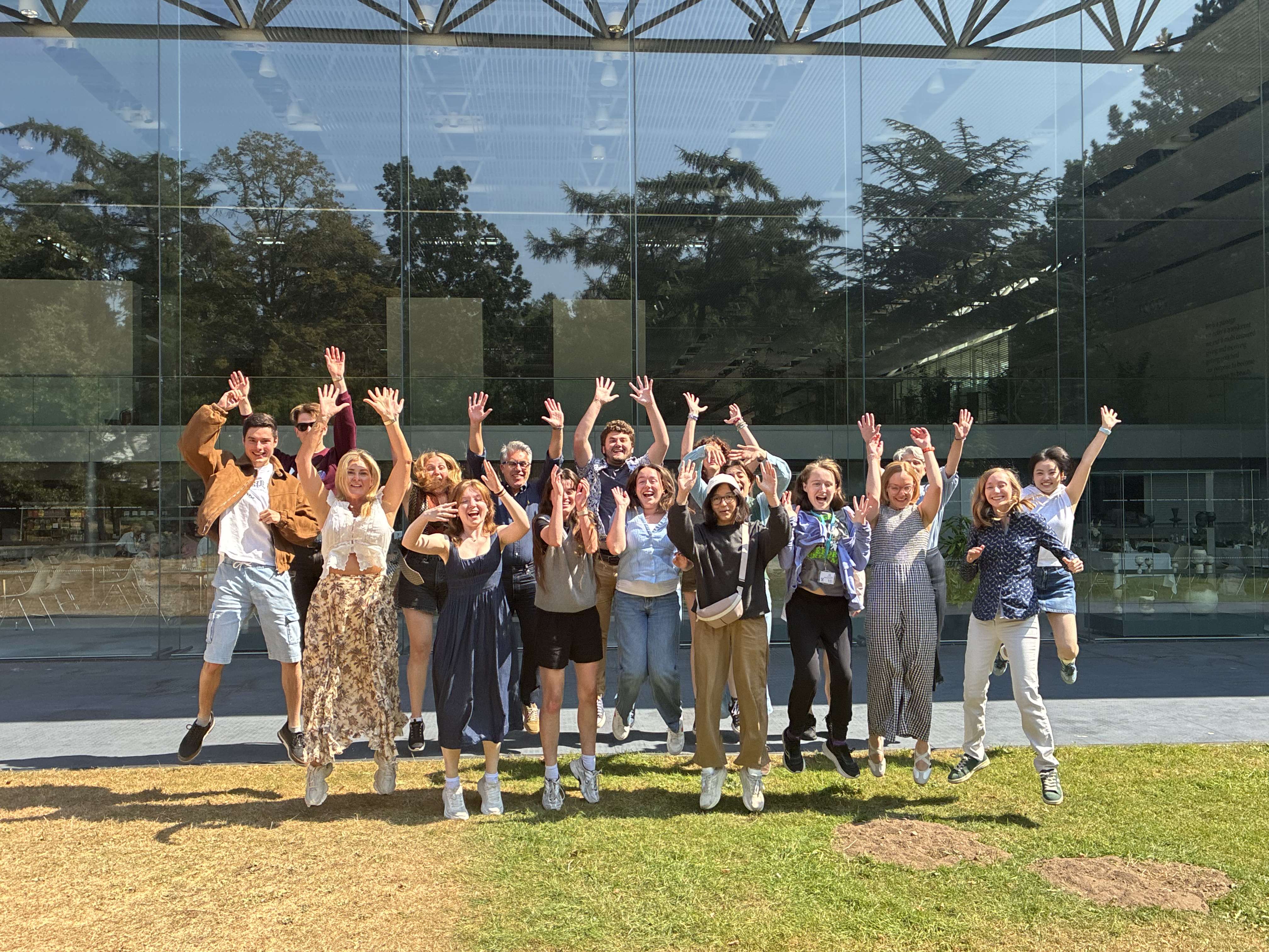 A group of students from the 2026 UEA Creative Writing Summer School cohort standing outside on the grass in front of the Sainsbury Centre cafe. They are standing in a big group and jumping in the air, with big smiles.