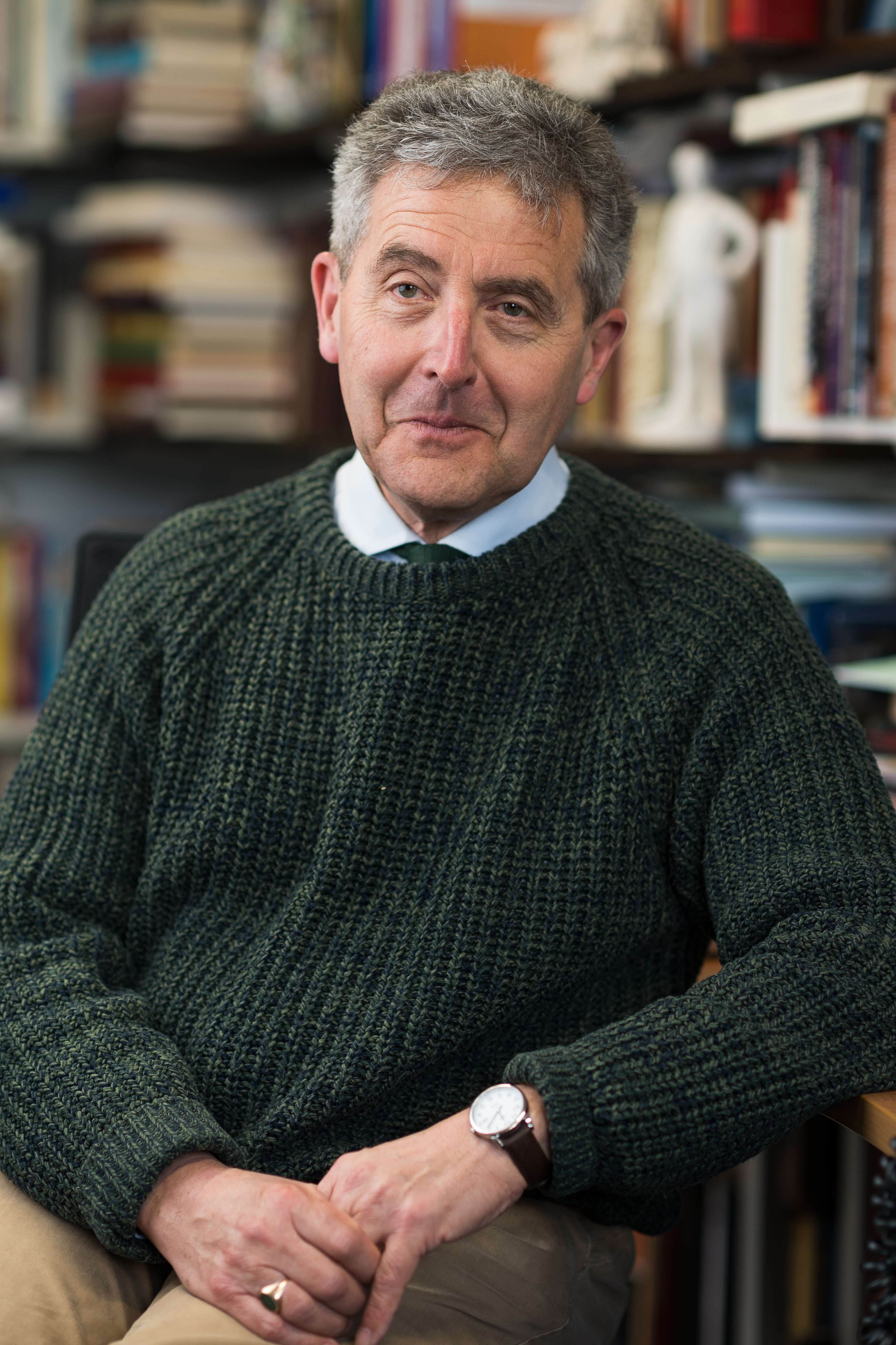 A portrait photo of a person in an office full of books. They are wearing a green jumper.
