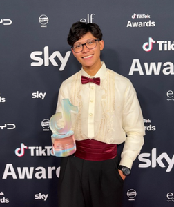 Gab Astorga, who is wearing glasses with a white shirt and red bow tie, is holding a TikTok award. He is standing in front of a black background with brand logos on it.