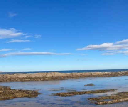 Rocky shoreline with calm water under a clear blue sky, scattered with a few clouds.