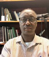 A person wearing glasses and a striped shirt is sitting in front of a bookshelf filled with various books and items.