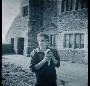 A smiling man wearing glasses holds a bird in his hands. He is standing in front of an old stone building with windows.