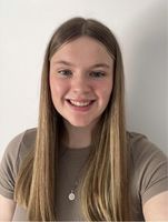 A young woman with long straight hair, wearing a brown top and a necklace, smiles at the camera against a plain white background.