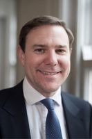 Smiling man in a suit and tie, standing indoors near a window with soft lighting.