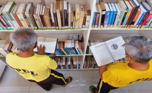 Two people in yellow shirts read books in front of a bookshelf filled with various books, seen from above.