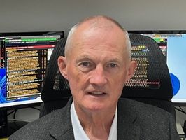 Man in a suit sitting in front of computer monitors displaying text and data, with a neutral expression.