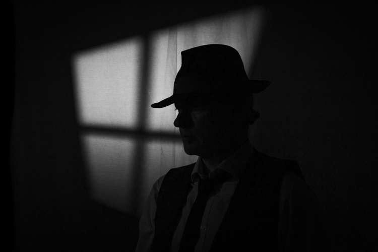 A black and white image showing the shadow of someone wearing a fedora. It is reminiscent of a film noir detective.