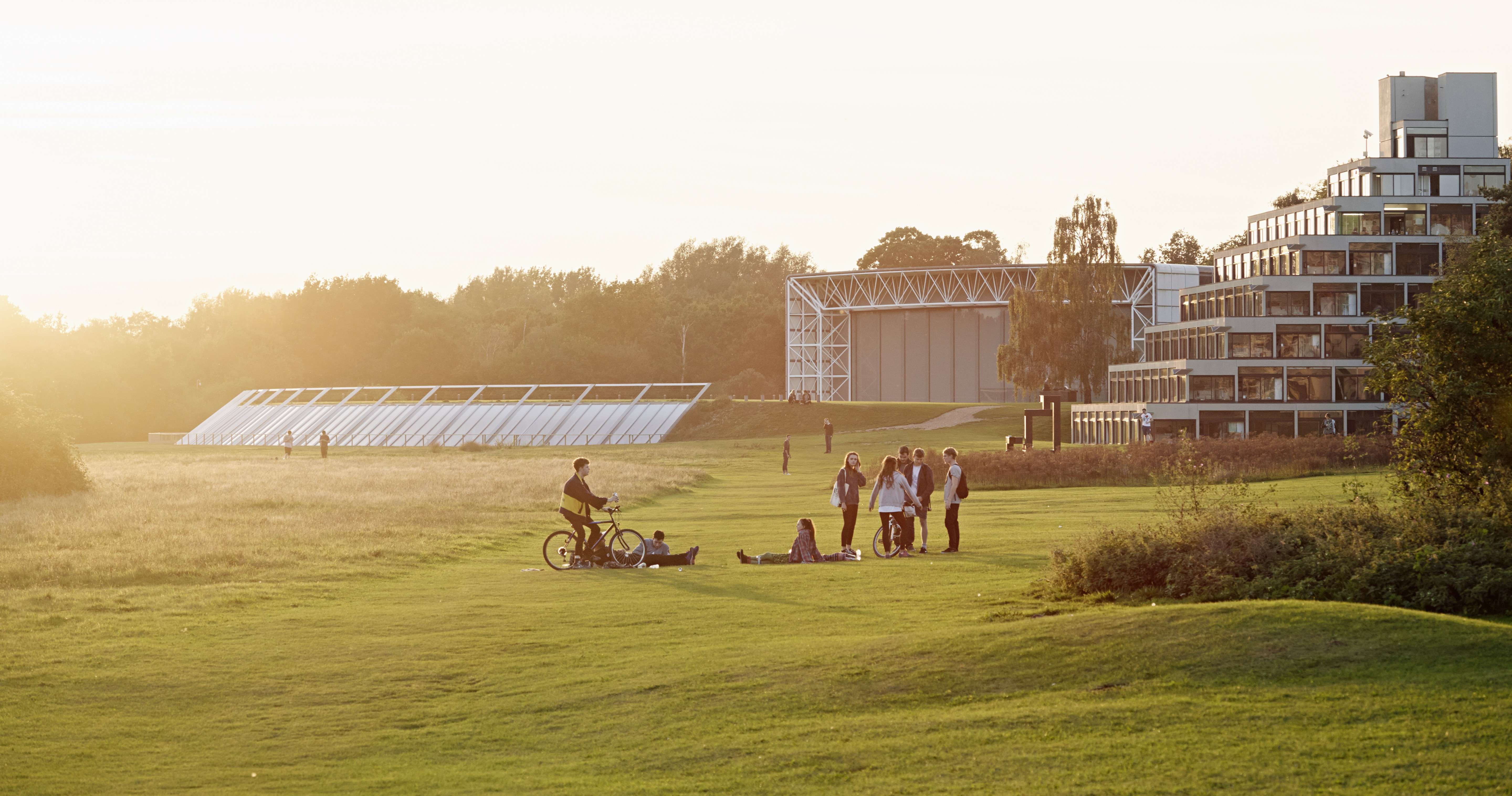 Several students standing and sitting on grass in front of the UEA Ziggurats and Sainsbury Centre at dusk.