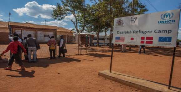 People entering Dzaleka Refugee Camp, with a sign displaying UNHCR and various national flags under a blue sky.