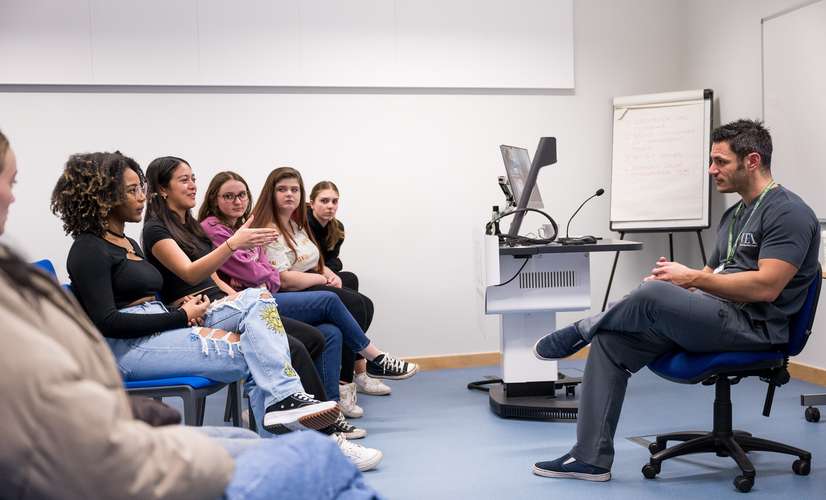 Group of students in small lecture room in discussion with health sciences lecturer wearing UEA branded scrubs