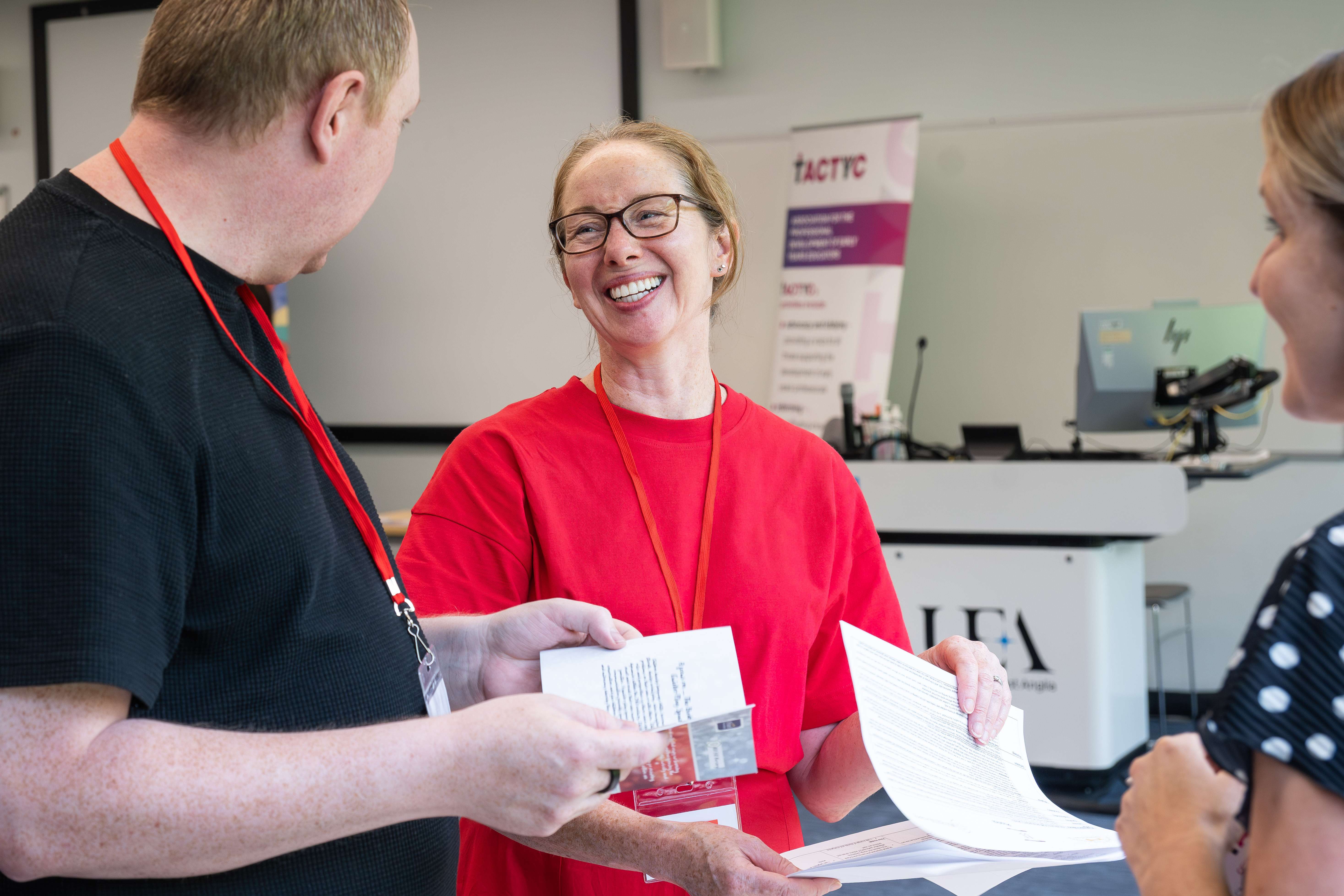 A smiling academic chats with attendees at a research conference, looking over papers and networking inside a modern university lecture hall.