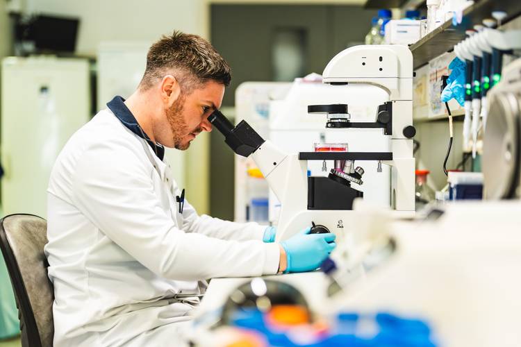 Dr Darrell Green, in a white lab coat and blue gloves, looks into a microscope in a laboratory setting surrounded by scientific equipment.