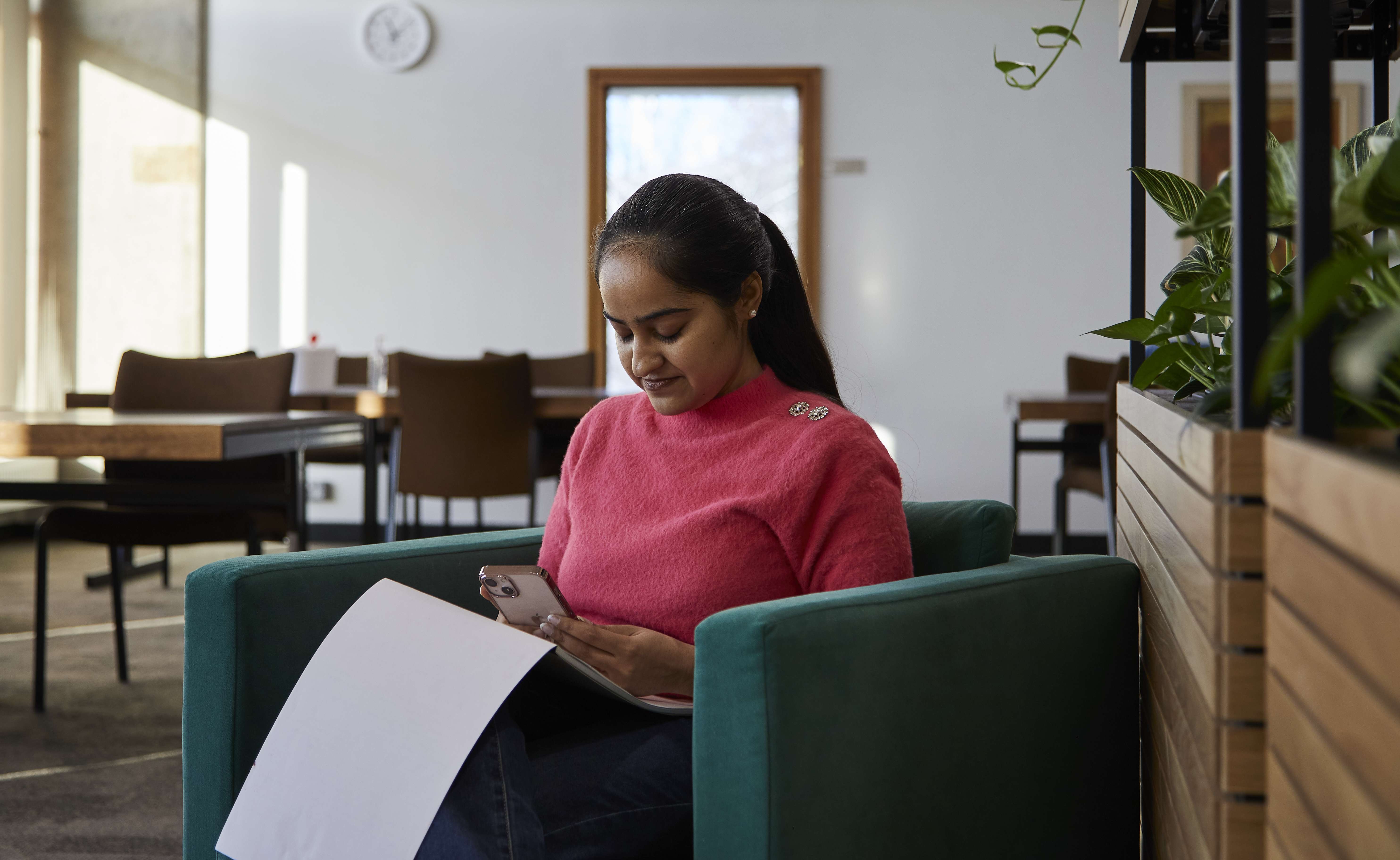 Person looking down at phone, on chair, plants next to them, tables and desks behind them