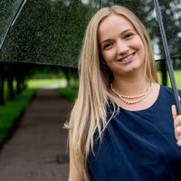 Ella, a young woman with long blonde hair wearing a pearl necklace and navy sleeveless top, smiling under an umbrella in nature