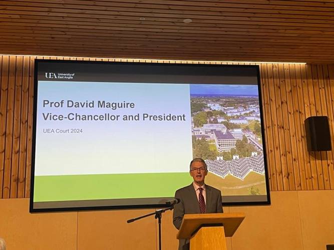 UEA Vice-Chancellor David Maguire speaking at a podium with a large screen behind him showing an image of UEA’s iconic architecture.