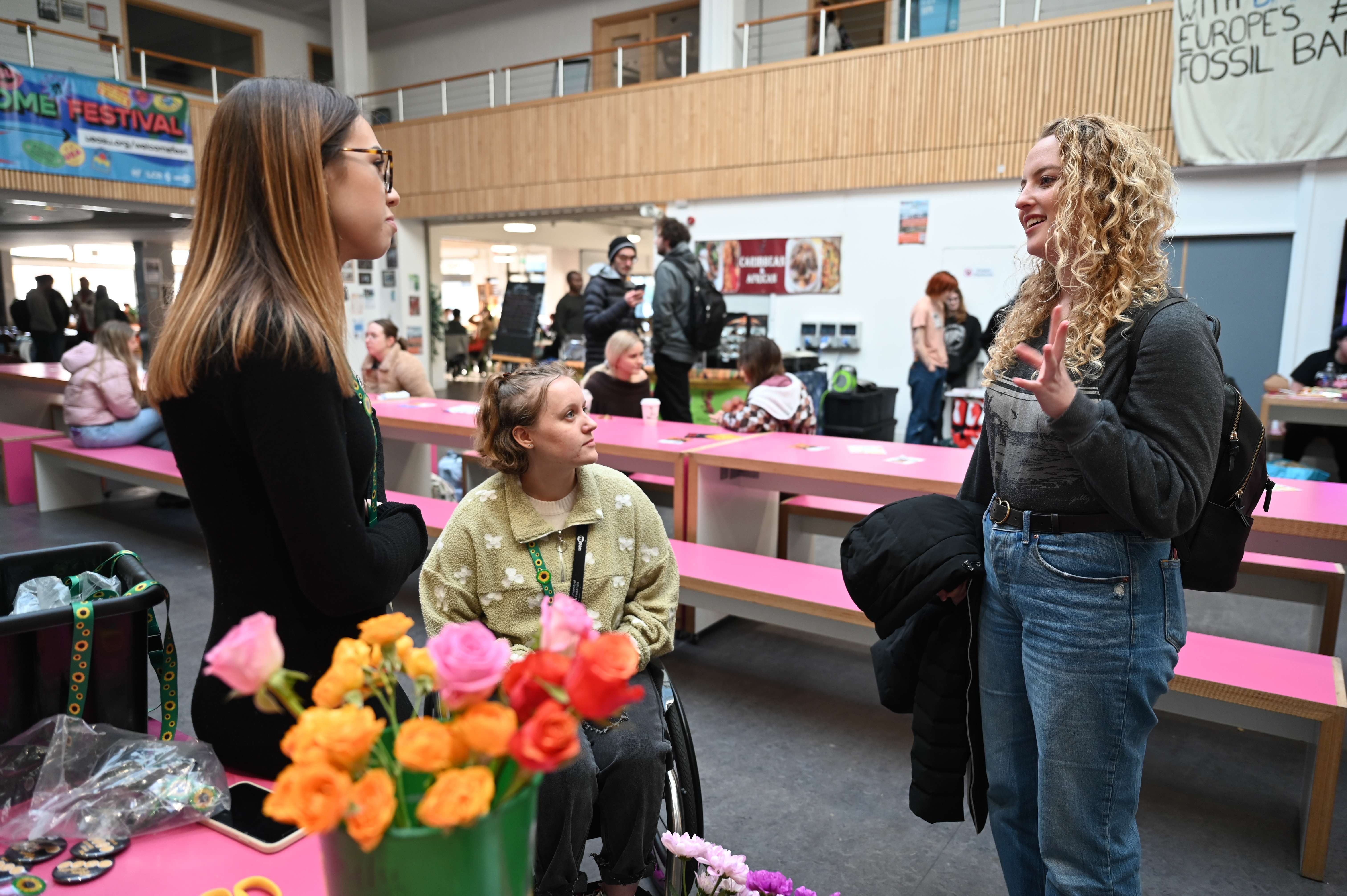 Three students together in Union House, one is in a wheelchair and two are standing either side, flowers in the foreground