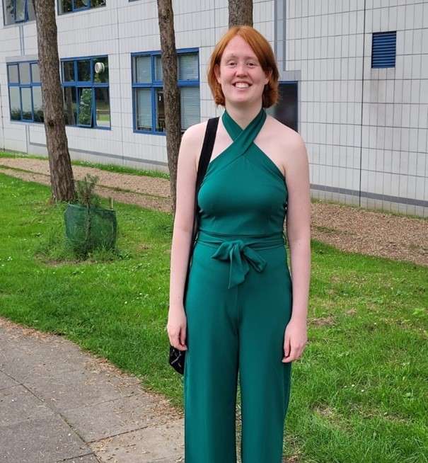 Meg in a green halter jumpsuit standing on a sidewalk, with grass, trees, and a building in the background.