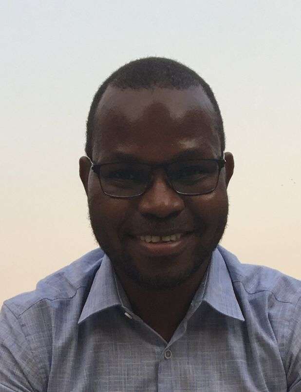 Smiling man wearing glasses and a light blue shirt against a clear sky backdrop.