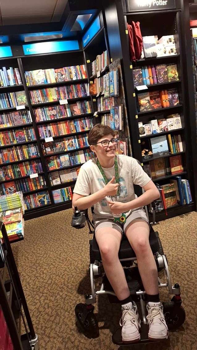 Osian in a wheelchair smiling and giving a thumbs-up in a bookstore, surrounded by colorful books and displays.