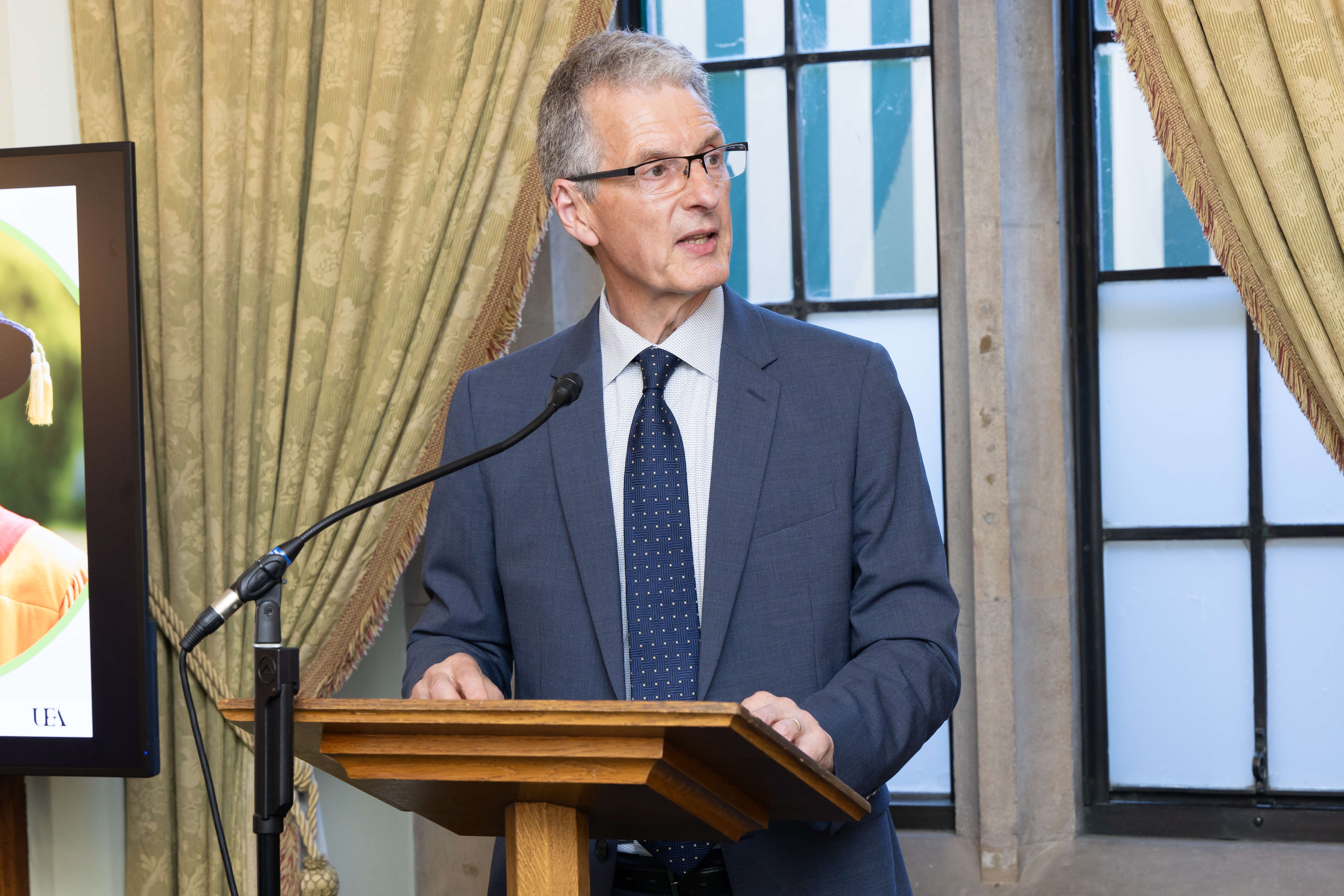 A man in a suit and glasses speaking at a wooden lectern by a curtained window.