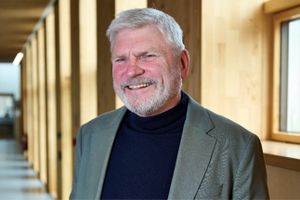Smiling man with grey hair and beard wearing a suit jacket and dark turtleneck, standing in a hallway with wooden walls and windows.
