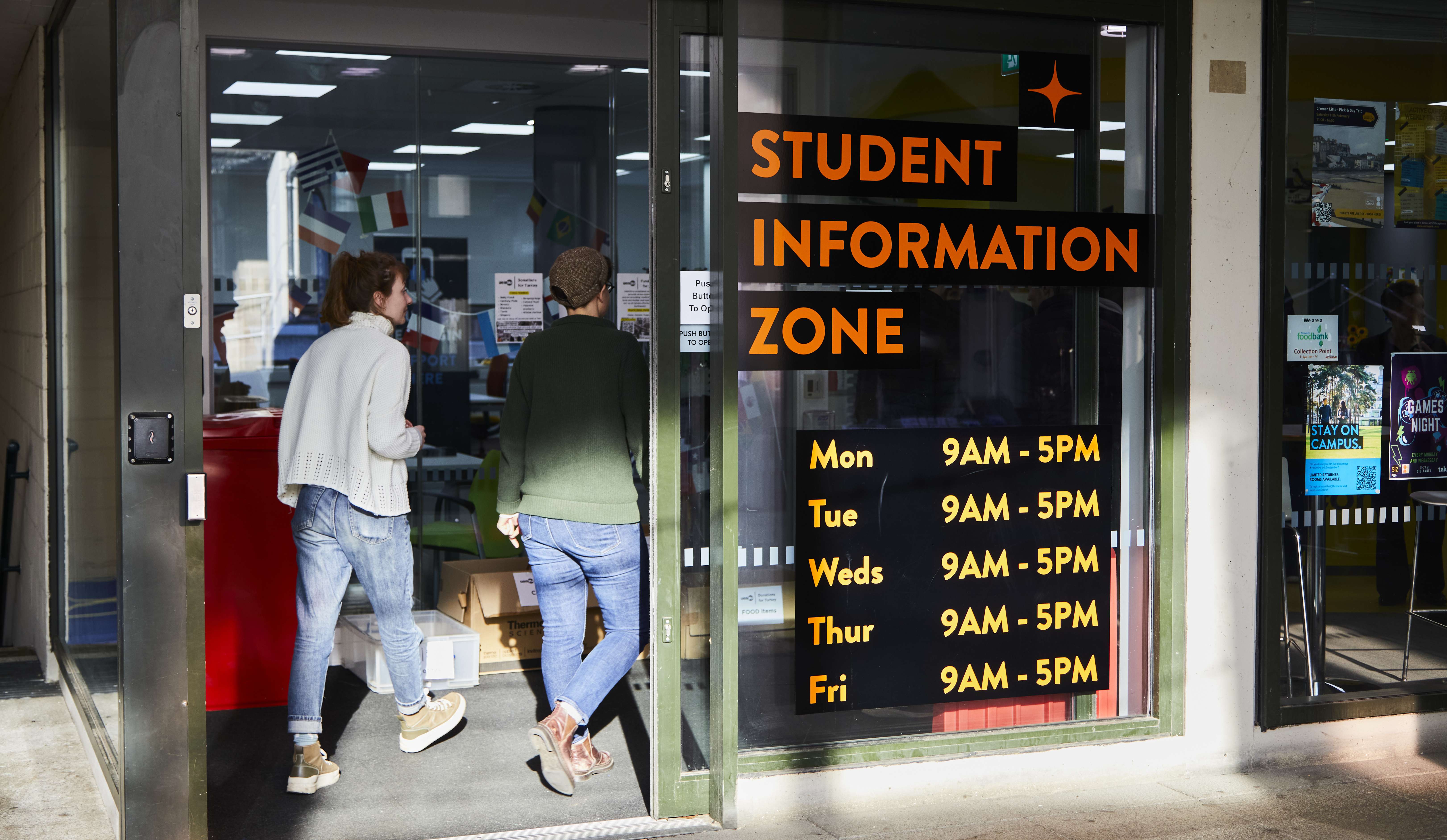 Two students walking in a door, on the window next to them says 'Student Information Zone' with opening times underneath