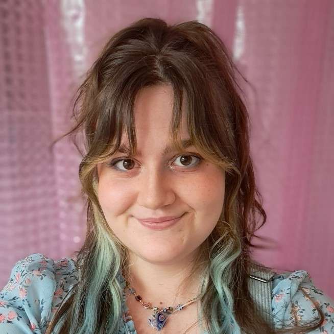 Libby smiling with long hair and blue highlights, wearing a floral top and necklace, stands in front of a pink background.