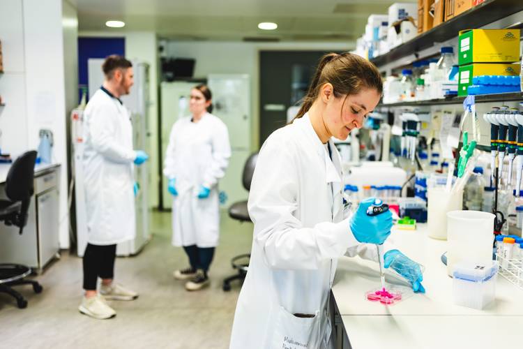 In a white lab coat and blue gloves, Emma Bull holds a pipette while standing in the foreground of the lab, focused on her work. In the background Dr Darrell Green and Amy Harden are conversing.