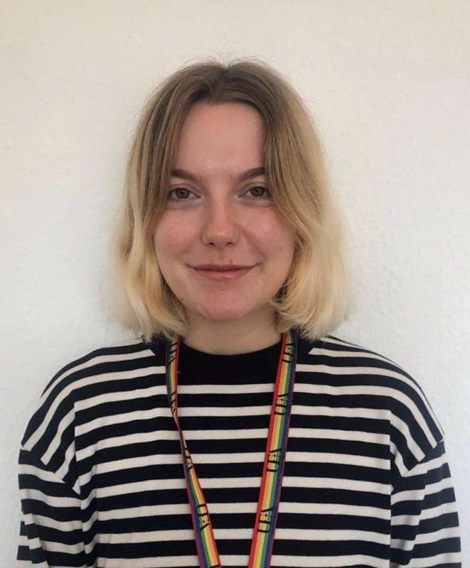 A person with short blonde hair wearing a black and white striped shirt and a rainbow lanyard stands against a white background.