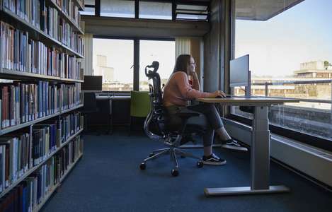 student at at a computer in the library with bookshelves behind her, sat on an adjustable chair