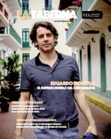 The cover of Issue 8 of the La Taberna magazine featuring a half-body image of a man wearing a short-sleeved collared shirt and jeans walking down a street with white buildings alongside and behind him