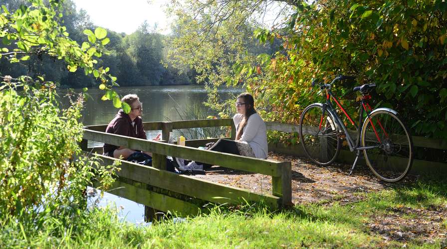 Two students talking and sitting in the sun by UEA lake surrounded by greenery and next to a red and black bike