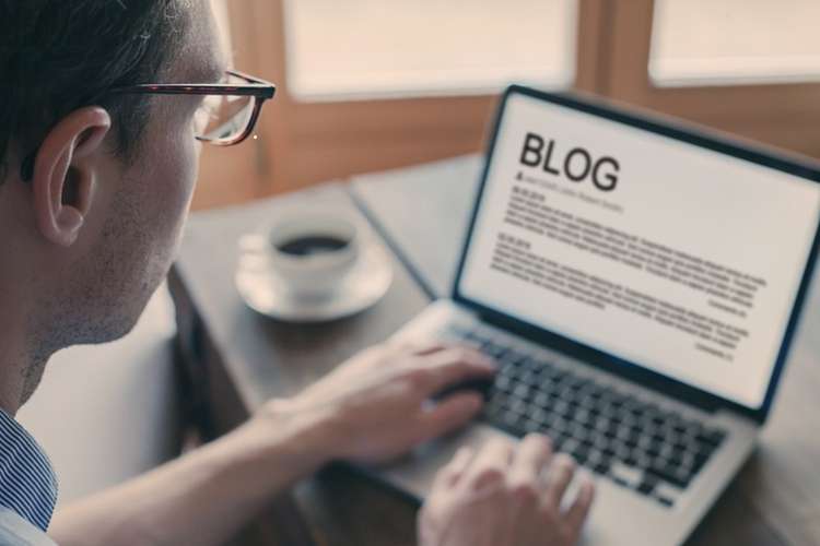 A postgraduate student with glasses writes a research blog post on their laptop, with a coffee on the desk.