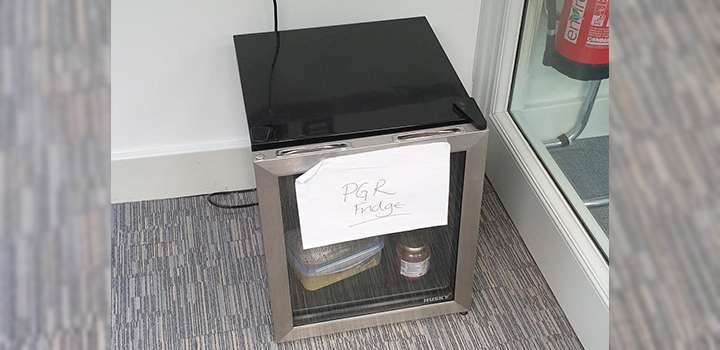 A small fridge on the floor with a white sheet of paper stuck to it with the words 'PGR Fridge'.