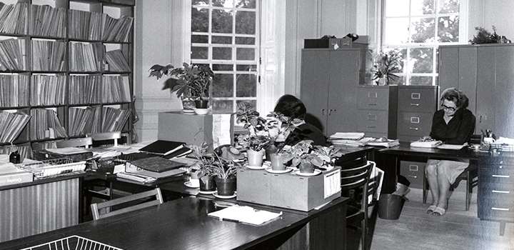 A black and white photo of people working at desks.