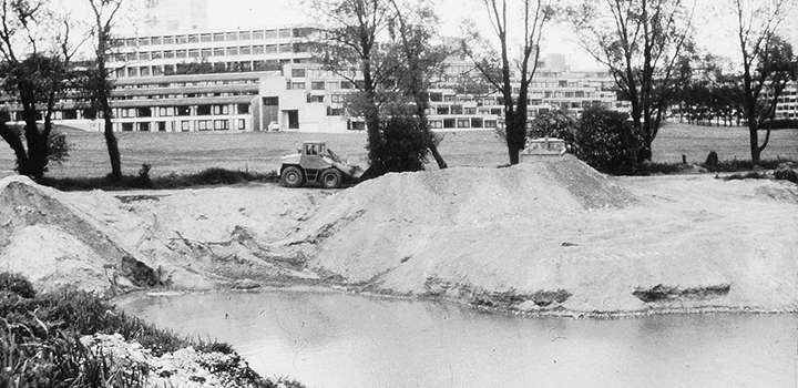 A black and white image of a machine working next to an earthwork by a lake.