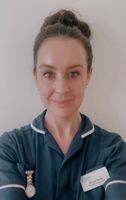 A woman in a blue nurse uniform with a name badge and a watch pin smiles at the camera against a plain background.