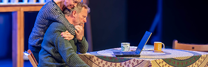 A standing woman hugs a man from behind as he sits at a table looking at a laptop. They look distressed.