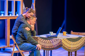 A standing woman hugs a man from behind as he sits at a table looking at a laptop. They look distressed.