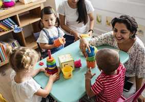 3 children sit around a table playing with toys. Two adults are playing with them.