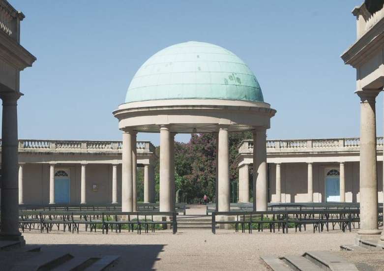 Neoclassical pavilion with a domed roof, surrounded by columns and symmetrical buildings, set in a courtyard under a clear blue sky.