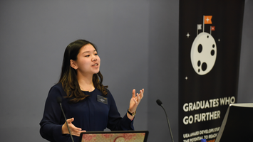 A woman in a blue blouse speaks at a podium. A banner reads "Graduates Who Go Further" with a moon and flags graphic.