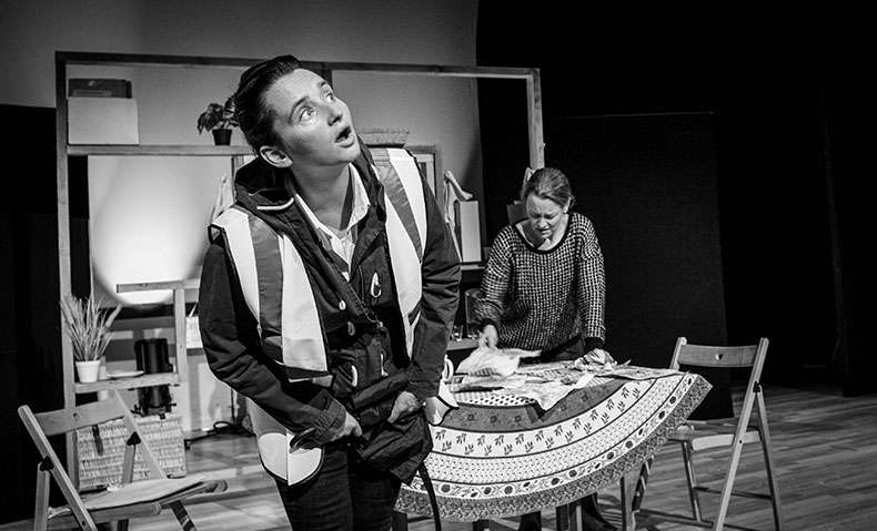 A woman in a high vis vest is in the foreground. In the background, another woman stands over a table and looks unhappy.