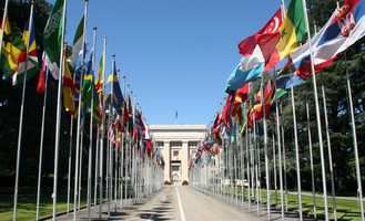 A wide pathway lined with numerous international flags leads to a large, stately building under a clear blue sky.