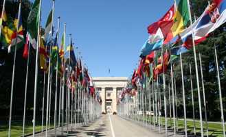 A wide pathway lined with numerous international flags leads to a large, stately building under a clear blue sky.