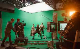 Film crew shoots actors in period costumes on a green screen set, with cameras and lighting equipment visible.