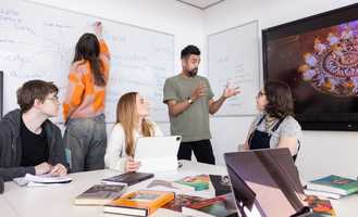 A group of students in a classroom, one writing on a whiteboard, others listening to a speaker, with books and a tablet on the table.