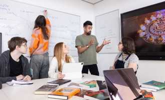 A group of students in a classroom, one writing on a whiteboard, others listening to a speaker, with books and a tablet on the table.