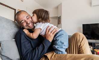 A child kisses a smiling man on the cheek as they sit together on a couch in a cozy living room.
