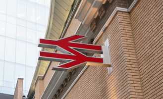 Red and silver angular logo mounted on a brick wall, with a glass building in the background.